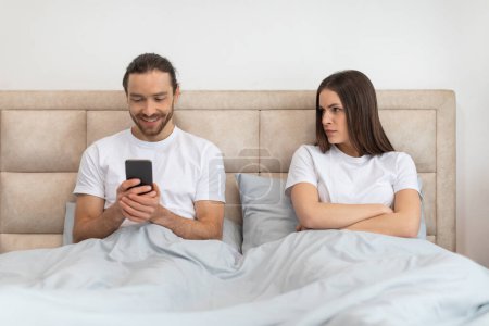 Téléchargez les photos : L'homme est heureux engagé avec son smartphone tandis que la femme à côté de lui regarde avec inquiétude et mécontentement l'expression, faisant allusion à la distraction numérique - en image libre de droit