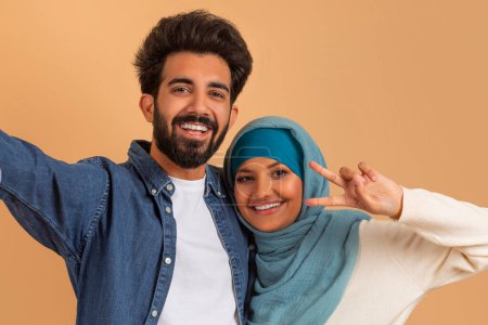 Téléchargez les photos : Joyeux jeune couple musulman prenant Selfie ensemble, joyeux époux arabes homme et femme dans Hijab posant et souriant à la caméra, debout ensemble sur fond de studio beige, gros plan - en image libre de droit