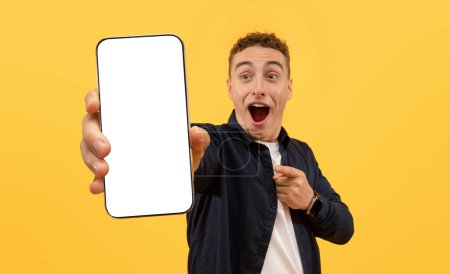 Téléchargez les photos : Choqué émotionnel jeune homme européen montrant grand smartphone avec écran blanc blanc maquette copie espace pour la publicité de l'application mobile, isolé sur fond jaune - en image libre de droit