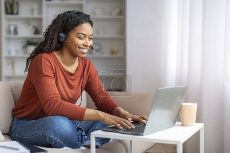 Photo for Smiling Young Black Woman Wearing Headset Using Laptop Computer At Home, Happy African American Female Sitting At Desk In Living Room, Enjoying Online Education And Distance Learning, Free Space - Royalty Free Image