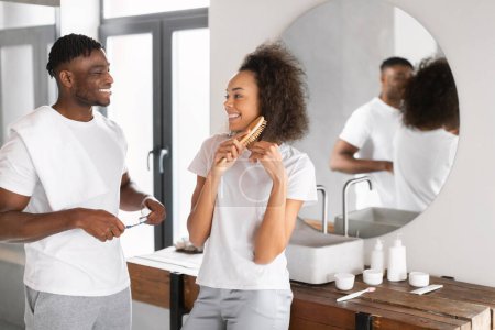 Glückliches schwarzes Paar bereitet sich gemütlich gemeinsam vor und verbringt Zeit im modernen Badezimmer, während die Frau Haare putzt und der Mann Zahnbürsten, miteinander redet und ihren Morgen genießt