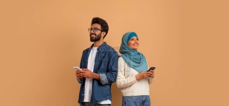 Photo for Modern lifestyle. Young arab couple standing back to back and holding smartphones, smiling muslim man and woman in hijab looking at copy space while standing on beige studio background, panorama - Royalty Free Image