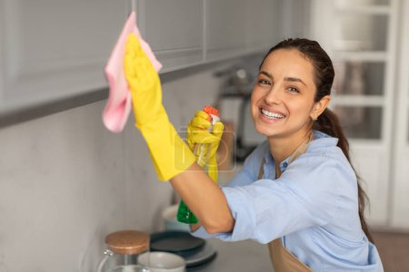 Foto de Mujer radiante en camisa y guantes amarillos limpia con entusiasmo la superficie de la cocina con botella de aerosol y tela rosa, que representa las tareas domésticas de rutina - Imagen libre de derechos