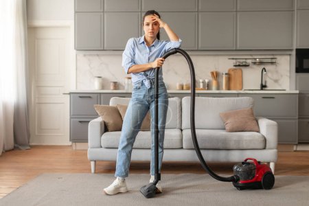 Photo for Tired young female in casual attire with vacuum cleaner standing in modern living room, showing fatigue from domestic chores against kitchen backdrop - Royalty Free Image