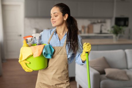 Photo for Contented woman in casual attire and yellow gloves carries bucket full of colorful cleaning products and cloths, standing in well-lit, modern kitchen, copy space - Royalty Free Image