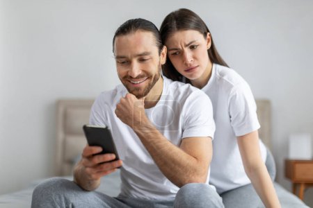 Foto de Mujer preocupada mirando por encima del hombro de los hombres sonrientes en su teléfono inteligente, lo que sugiere secreto o curiosidad en el entorno de la habitación doméstica - Imagen libre de derechos
