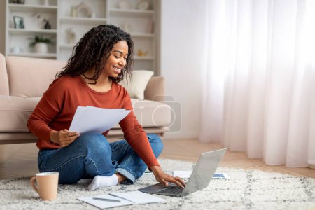 Téléchargez les photos : Femme indépendante noire souriante travaillant avec un ordinateur portable et des papiers à la maison, heureuse jeune femme afro-américaine assise sur le sol dans la chambre, vérifiant les documents et utilisant l'ordinateur, appréciant le travail à distance - en image libre de droit