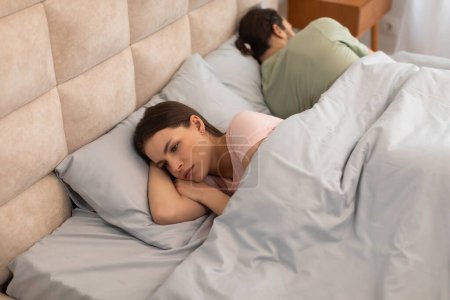 Upset young woman lies in bed turned away from her partner, capturing moment of emotional distance and tension in relationship within bedroom setting