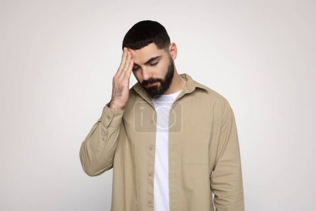 Photo for Man with a beard feeling stressed or having a headache, holding his forehead with a pained expression, dressed in a beige shirt and white tee, on a soft background, studio - Royalty Free Image