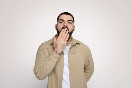 Téléchargez les photos : Fatigué homme arabe millénaire avec une barbe couvrant sa bouche avec la main, portant une veste beige sur une chemise blanche, exprimant choc ou incrédulité sur un fond uni, studio - en image libre de droit