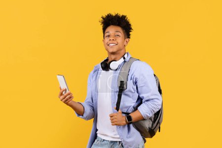 Téléchargez les photos : Étudiant masculin ravi avec sac à dos, écouteurs autour du cou, tenant smartphone, incarnant l'ambiance de l'éducation à l'âge numérique sur fond jaune vif - en image libre de droit