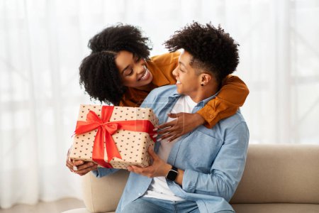 Photo for A young, cheerful African American couple sharing a moment of joy while exchanging a heart-patterned gift box with a vibrant red ribbon in a cozy living room setting at home - Royalty Free Image