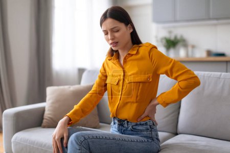 Junge Frau in lässiger Kleidung fühlt sich unwohl, umklammert ihren unteren Rücken, während sie auf einer modernen bequemen Couch sitzt und unter akuten Rückenschmerzen leidet