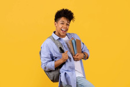 Foto de Un animado estudiante negro con una sonrisa encantadora, con una mochila y cuadernos que se aferran, exuda entusiasmo y preparación para un nuevo día académico contra un vibrante fondo amarillo. - Imagen libre de derechos