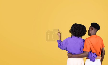 Téléchargez les photos : Vue arrière d'un jeune couple debout ensemble, la femme en chemisier violet pointant quelque chose au loin tandis que l'homme en chemise orange regarde, sur un fond jaune uni - en image libre de droit