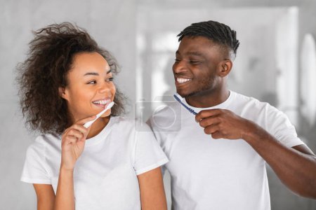 Téléchargez les photos : Soins dentaires. Romantique couple afro-américain brossant les dents ensemble debout côte à côte. Hygiène buccale familiale dans la salle de bain moderne à l'intérieur, conjoints posant avec des brosses à dents souriant à la caméra - en image libre de droit