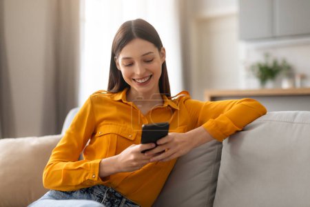 Foto de Mujer joven y relajada en blusa disfruta navegando en su teléfono inteligente, descansando cómodamente en un sofá gris con luz natural iluminando su sonrisa contenta - Imagen libre de derechos