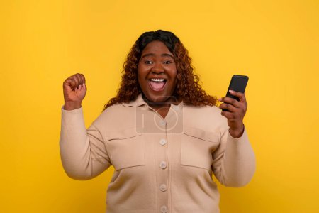 Photo for Great online offer. Emotional long-haired young chubby african american lady with smartphone in her hand gesturing and exclaiming, yellow studio background. Bet on Internet, easy cash - Royalty Free Image
