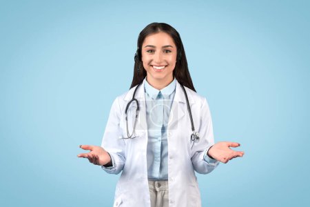 Photo for Approachable female doctor with friendly smile, open hands gesturing welcome, wearing white lab coat and stethoscope and smiling, against blue background - Royalty Free Image