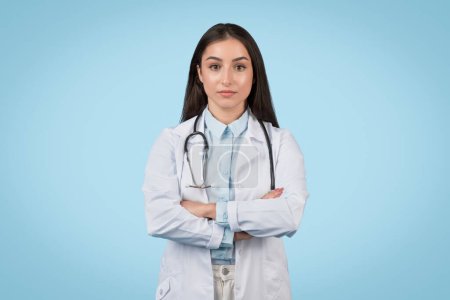 Téléchargez les photos : Jeune médecin professionnelle portant un manteau blanc et un stéthoscope, les bras croisés, debout en toute confiance sur un fond bleu solide - en image libre de droit