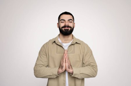 Foto de Un hombre árabe milenario con los ojos cerrados y una sonrisa suave muestra un gesto namaste, vestido con una chaqueta beige casual sobre una camiseta blanca, contra un telón de fondo neutral, estudio - Imagen libre de derechos