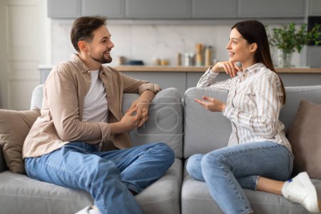 Smiling young man and woman sit comfortably on gray sofa, deeply involved in cheerful and animated conversation, exemplifying joyful domestic moment