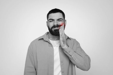 Photo for Black and white image of millennial arab man with a beard feeling dental discomfort, holding his cheek with a pained expression, highlighted by a red spot on his jaw, suggesting tooth pain - Royalty Free Image