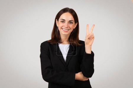 Foto de Joven mujer caucásica positiva en un traje negro profesional chaqueta gestos signo de paz con una sonrisa amistosa, de pie con confianza en un fondo gris claro, estudio. Trabajo, negocios - Imagen libre de derechos