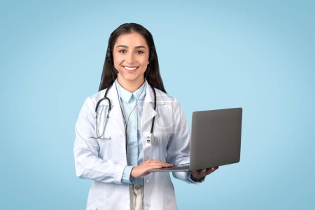 Photo for Professional female doctor with headset using laptop, showcasing multitasking ability and online consultation, dressed in lab coat on calming blue background - Royalty Free Image