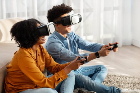 Téléchargez les photos : Couple afro-américain millénaire enchanté plongé dans un jeu de réalité virtuelle, portant des casques VR et tenant des contrôleurs, assis sur un tapis doux dans un salon ensoleillé à la maison - en image libre de droit