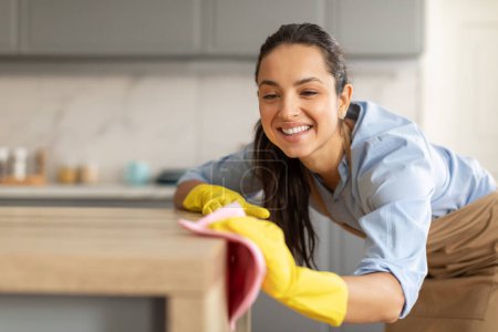 Foto de Mujer joven radiante en camisa azul y guantes amarillos, limpiando el polvo de la mesa de madera con sonrisa, mostrando limpieza de rutina en la cocina brillante, espacio libre - Imagen libre de derechos