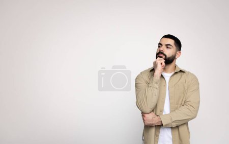 Téléchargez les photos : Pensif homme arabe millénaire avec une barbe dans une veste beige et chemise blanche tient menton, contemplant, avec une expression réfléchie sur un simple, regarder l'espace libre, fond propre, studio - en image libre de droit
