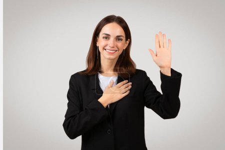 Photo for Pleasant millennial caucasian businesswoman with a sincere smile raises her right hand to take an oath or pledge, placing her left hand over her heart, signifying honesty and commitment - Royalty Free Image