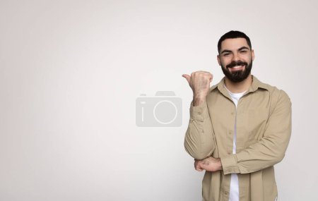 Foto de Alegre hombre barbudo milenario árabe dando un signo de pulgar hacia arriba, sonriendo con confianza en una camisa beige casual con mangas enrolladas y una camiseta blanca, de pie sobre un fondo claro - Imagen libre de derechos