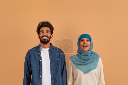 Foto de Retrato de pareja musulmana emocionada mirando hacia arriba en el espacio de copia por encima de sus cabezas con la boca abierta, hombre y mujer árabe sorprendido en hijab impresionado por la buena oferta, de pie juntos sobre fondo beige - Imagen libre de derechos