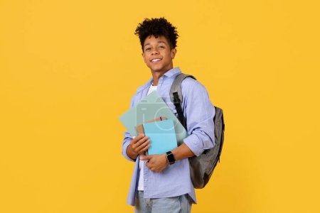 Téléchargez les photos : Un étudiant noir joyeux avec une coupe de cheveux élégante et un sac à dos retient avec confiance les manuels, prêt pour la journée d'apprentissage sur un fond jaune vibrant - en image libre de droit