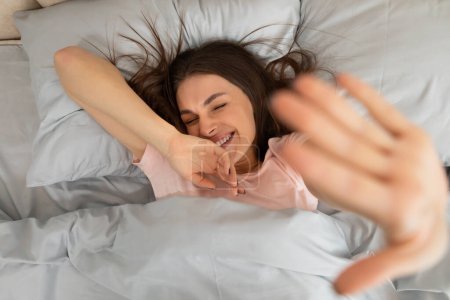 Téléchargez les photos : Femme heureuse se réveillant, étendant la main à la caméra et souriant, couchée sur le lit dans la chambre, bonjour à la maison, profiter du week-end et du temps libre, vue sur le dessus - en image libre de droit