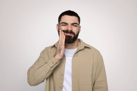 Photo for Man with a beard winces in discomfort, touching his cheek with a hand, suggesting dental pain or toothache, wearing a beige shirt and white t-shirt, with a light background - Royalty Free Image