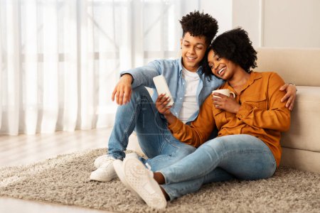 Photo for A cozy young african american couple in casual wear, comfortably relaxing on a plush rug, sharing a smartphone screen and enjoying a warm, intimate moment in their luminous living room - Royalty Free Image