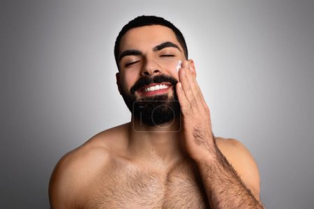 Téléchargez les photos : Joyeux beau jeune homme arabe barbu en utilisant sa crème visage préférée après la douche, posant torse nu sur fond de studio gris. Moyen-Orient homme appliquant crème solaire lotion - en image libre de droit