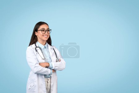 Photo for Upbeat female doctor wearing glasses and lab coat with stethoscope, arms crossed, gazing to side at free space with hopeful expression on blue background - Royalty Free Image