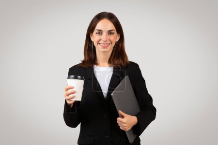 Foto de Sonriente mujer de negocios caucásica milenaria en una chaqueta negra sosteniendo una taza de café y una tableta desechables, listo para un día ocupado en el trabajo con todos los gadgets modernos esenciales - Imagen libre de derechos