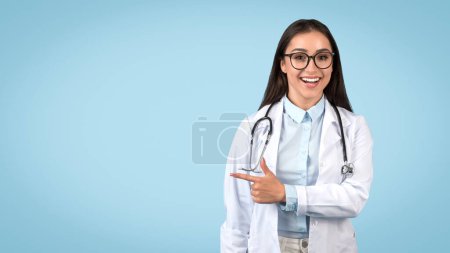 Foto de Médico de aspecto inteligente con gafas apuntando al espacio libre y sonriendo, en bata blanca y con estetoscopio, sobre fondo azul suave, panorama - Imagen libre de derechos