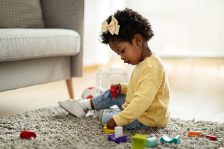 Téléchargez les photos : Adorable bébé afro-américain heureux fille jouant avec des jouets pour enfants à la maison tout en étant assis sur le sol de tapis dans le salon. Portrait de sourire mignon tout-petit en utilisant des jouets colorés, espace de copie - en image libre de droit