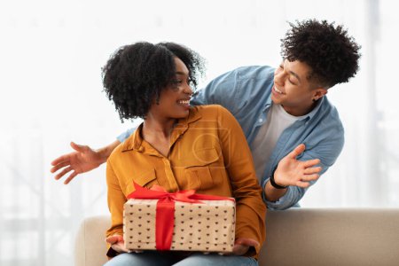 Photo for Excited African American millennial woman receiving a gift with heart patterns from a smiling man, creating a joyful and intimate moment of giving and surprise in a well-lit room - Royalty Free Image