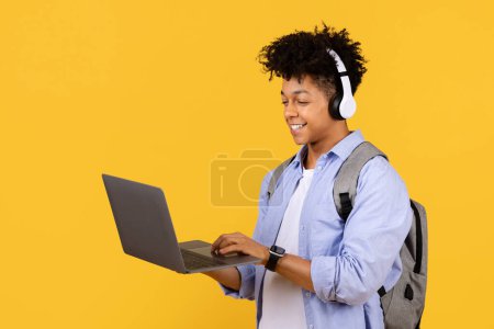 Foto de Alegre estudiante masculino negro equipado con auriculares profundiza en el aprendizaje interactivo en el ordenador portátil, destacando mezcla perfecta de ocio y estudio contra el fondo amarillo vibrante - Imagen libre de derechos
