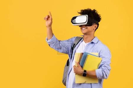 Photo for Cheerful black male student wears virtual reality headset, gesturing upwards as he holds notebooks, merging cutting-edge technology with traditional learning against vibrant yellow background - Royalty Free Image