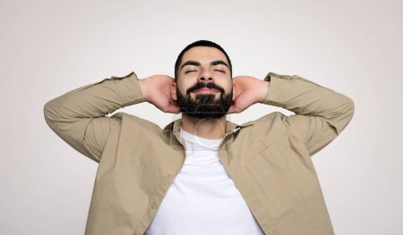 Téléchargez les photos : Un homme arabe millénaire détendu avec une barbe et un sourire content se penche en arrière avec les mains derrière la tête, portant une veste beige sur une chemise blanche sur un fond uni - en image libre de droit