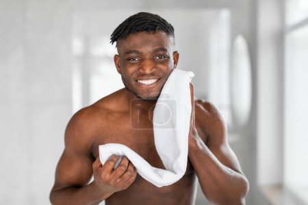 Téléchargez les photos : Portrait de joyeux homme millénariste noir essuie son visage avec une serviette dans la salle de bain moderne, debout torse nu et souriant à la caméra. Concept masculin de soins de la peau et de toilettage masculin - en image libre de droit