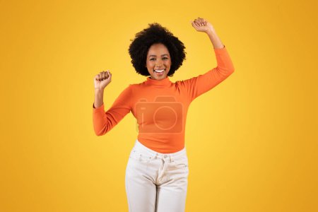 Foto de Sonriente joven afroamericana alza alegremente los brazos en un gesto de celebración, vistiendo un cuello alto naranja y vaqueros blancos sobre un fondo amarillo soleado, estudio - Imagen libre de derechos